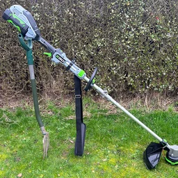 The Ego ST1511E-T grass trimmer propped upright with a spade, on a lawn in front of a hedge