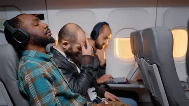 Three men on an aeroplane, two of whom are wearing wireless headphones