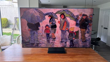 The Samsung R95H Micro LED TV on a wooden table in a conservatory displaying digital artwork of people with umbrellas