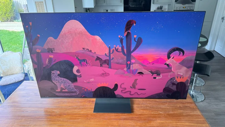 Top-down angled view of the Samsung R95H Micro LED TV on a wooden table in a conservatory displaying a digital artwork in the desert with various creatures