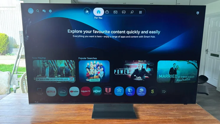 The Tizen OS home page being displaying on the Samsung R95H Micro LED TV on a wooden table in a conservatory