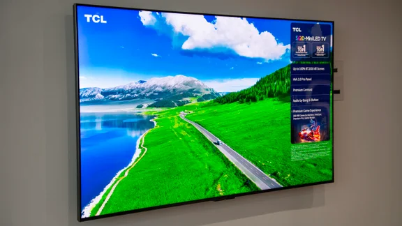 The TCL C7L Super QLED TV mounted on a wall displaying a scene featuring a snow-capped mountain, lake and green field bisected by a road
