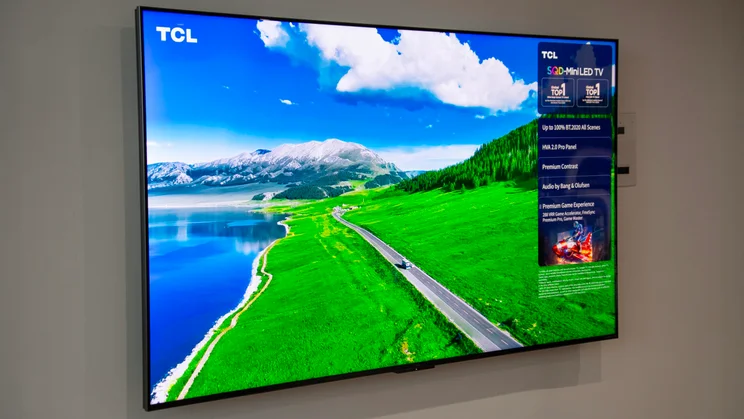 The TCL C7L Super QLED TV mounted on a wall displaying a scene featuring a snow-capped mountain, lake and green field bisected by a road