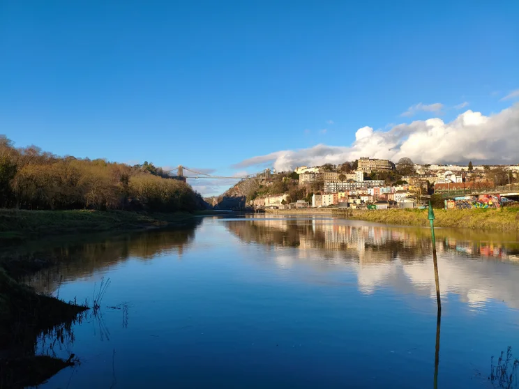 A wide river on a sunny day with a bridge crossing between two hills in the distance