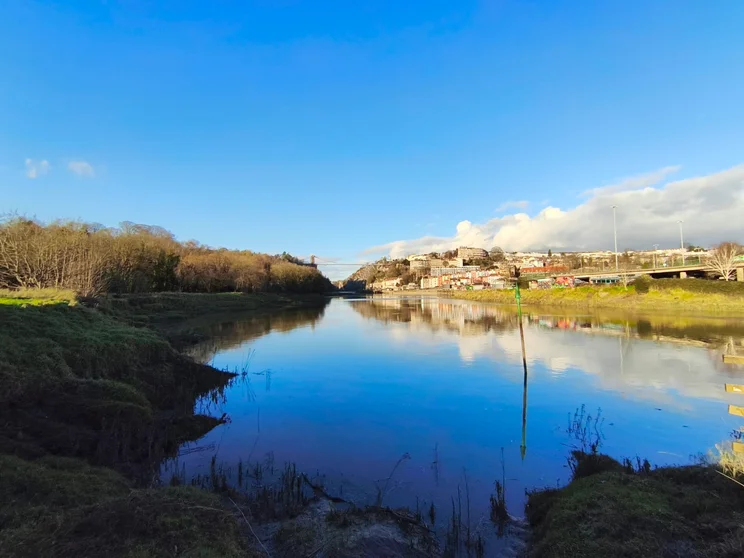 Ultrawide shot of a wide river on a sunny day, a bridge crossing between two hills in the distance